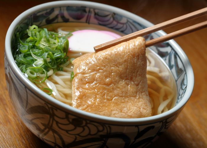 Kitsune udon, with udon, fishcakes, spring onion, and fried tofu in a bowl.